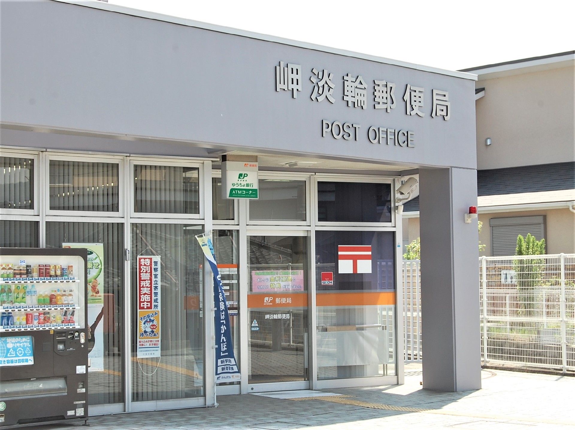 グレーの外壁の郵便局がある。特別警戒実施中の張り紙があるので、安心して利用することができる。自動販売機もある。