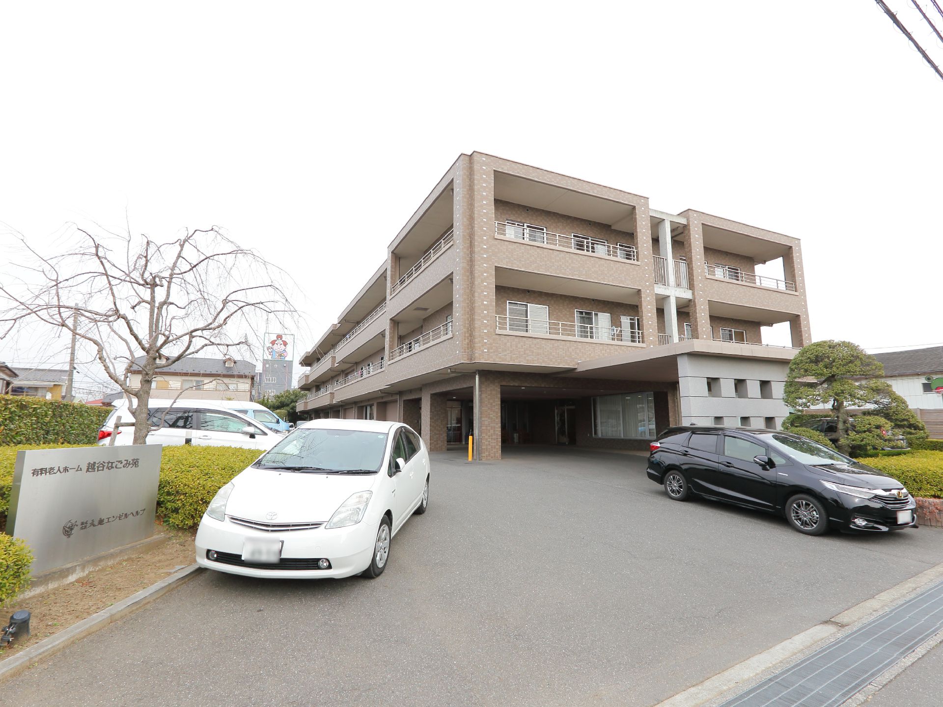 完全個室スタイルの老人ホームの外観で、駐車場が施設正面に設置されている。隣の建物との境界には、緑の生け垣がある。