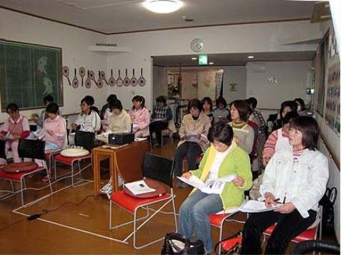 フローリングの部屋の中で揃いの赤い椅子に座り、集まっている大勢のスタッフたち