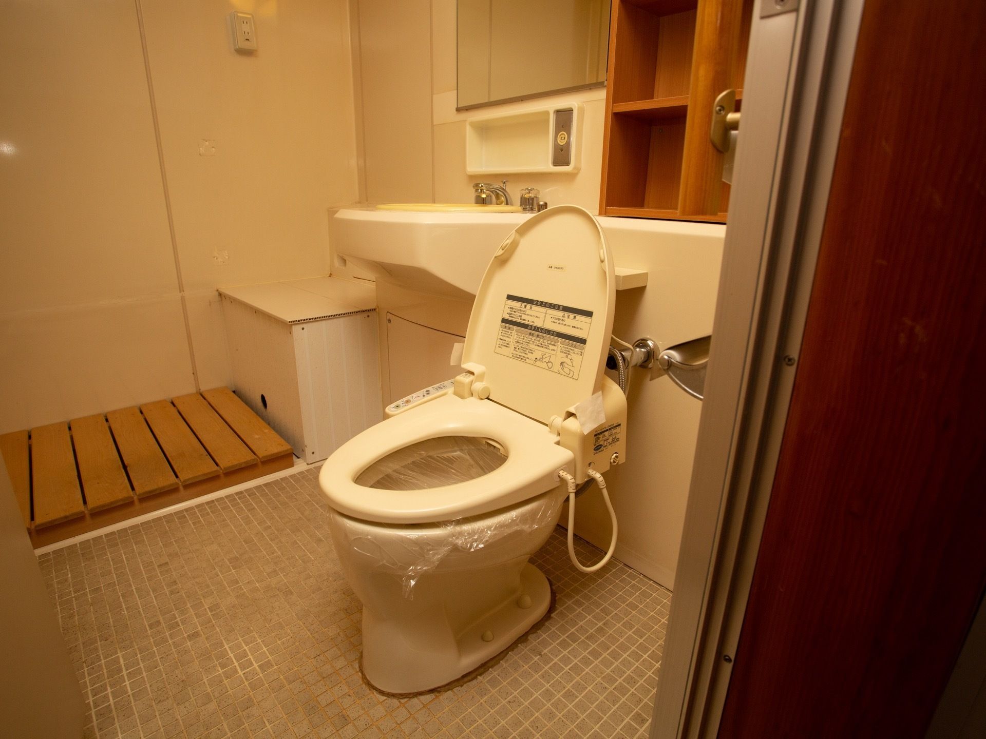 洗面台の横に温水洗浄便座器が完備されたトイレである。蓋は開けられ、便器内にはビニールが敷かれている。