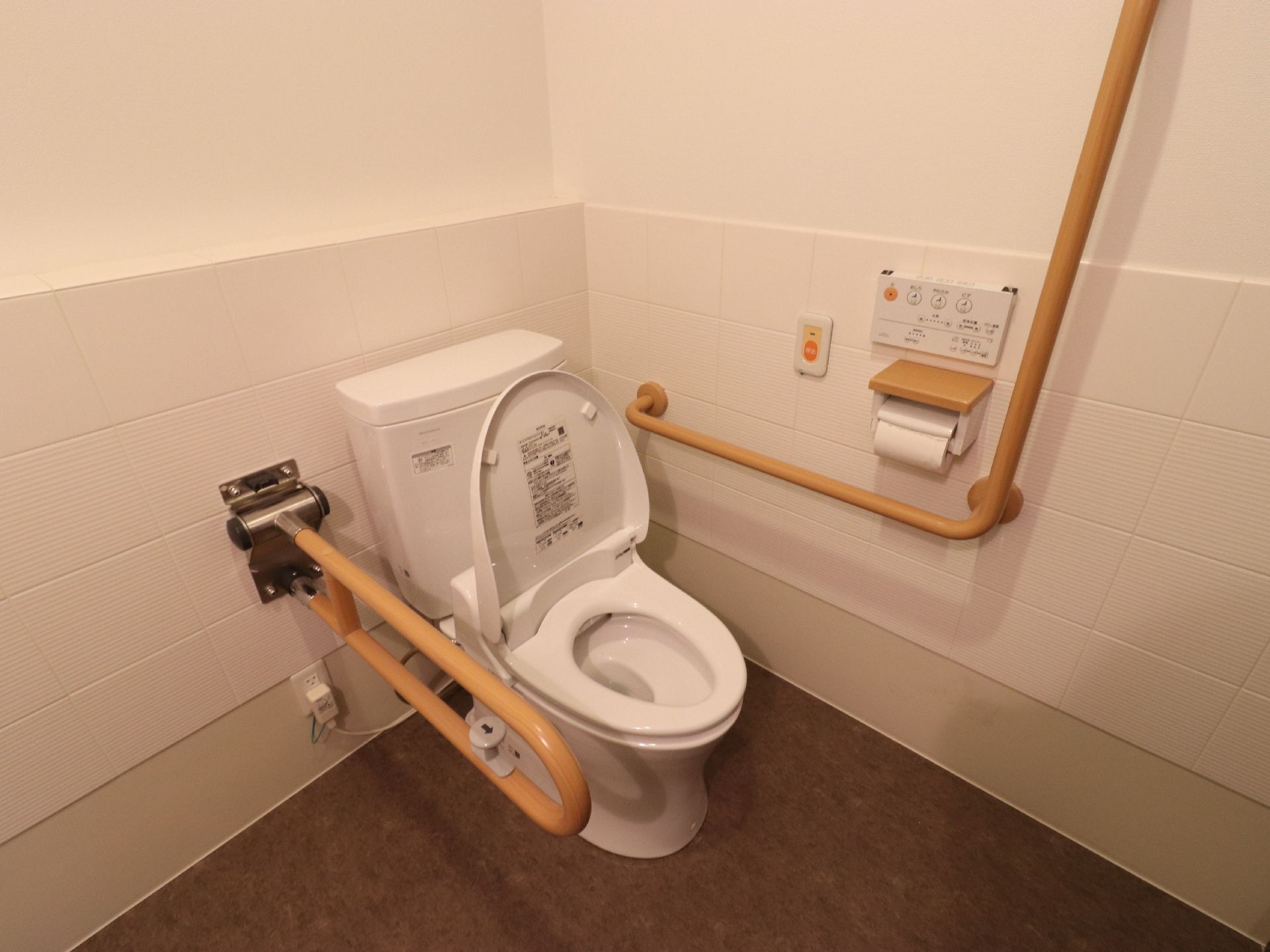 トイレには洋式便器が設置されている。便器の両側には形状の異なる手すりがある。壁側には操作パネルなどがついている。