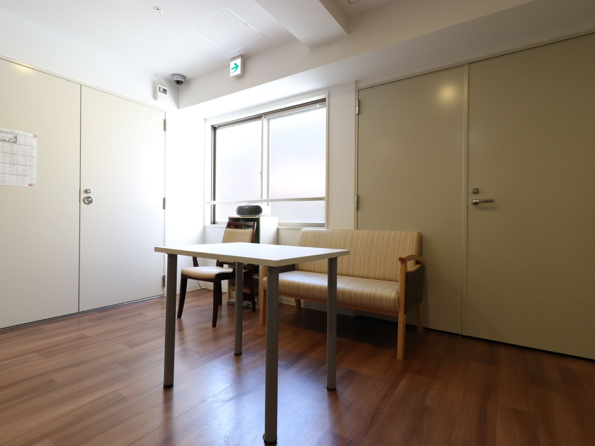 部屋の内側に柵が付いた窓がある。床はフローリングで、テーブルや椅子、ソファーが置かれている。白い扉には紙が1枚貼られている。