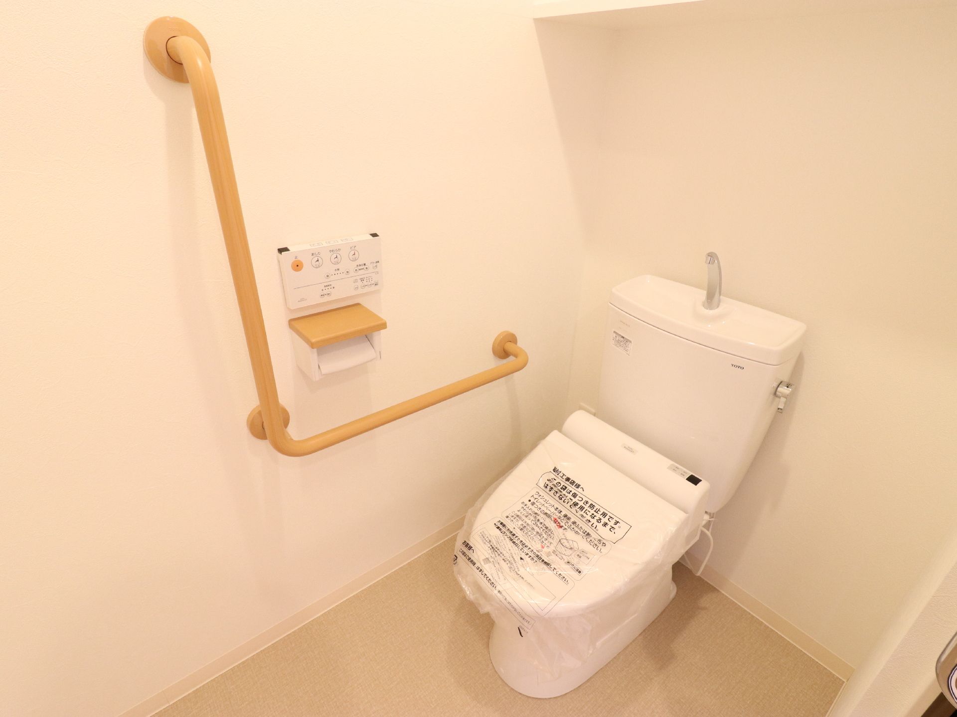 白い壁とベージュの床の空間に洋式便器が設置されている。壁側には手すりが付いている。トイレットペーパーフォルダーの上には操作パネルがある。