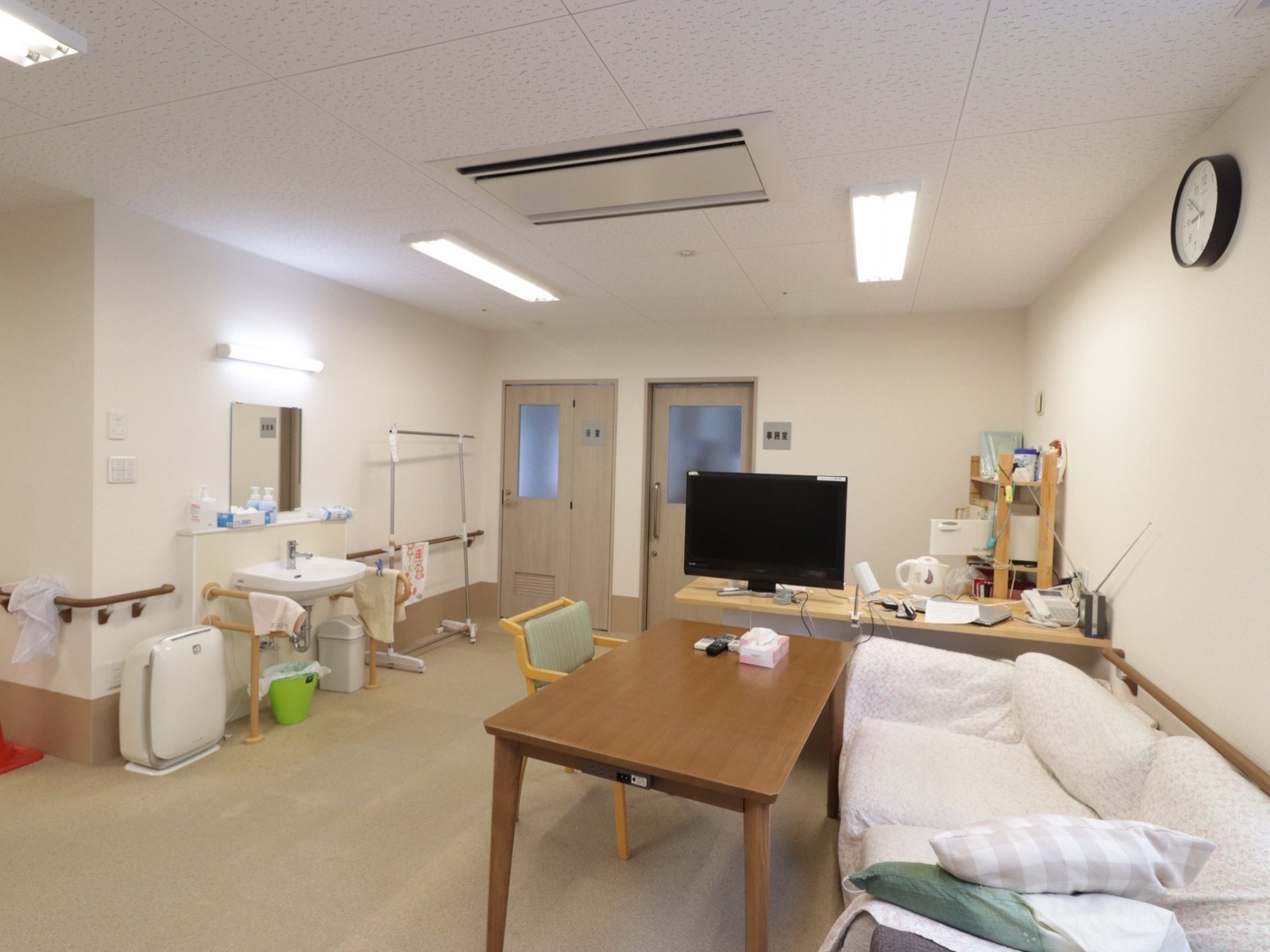 事務室の前のホールには、洗面台や空気清浄機が置かれている。テレビの前にはテーブルやカバーが掛かったソファーがある。