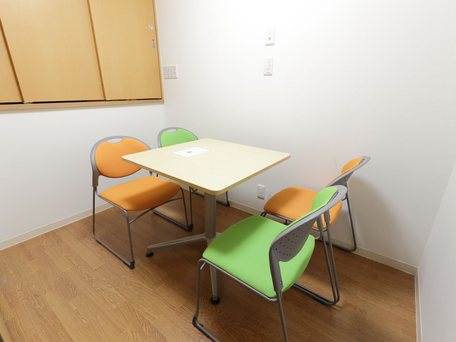 白い壁面の部屋に小さなテーブルが置いてある。オレンジと緑の椅子が4脚向かい合う形で置かれており床はフローリングになっている。