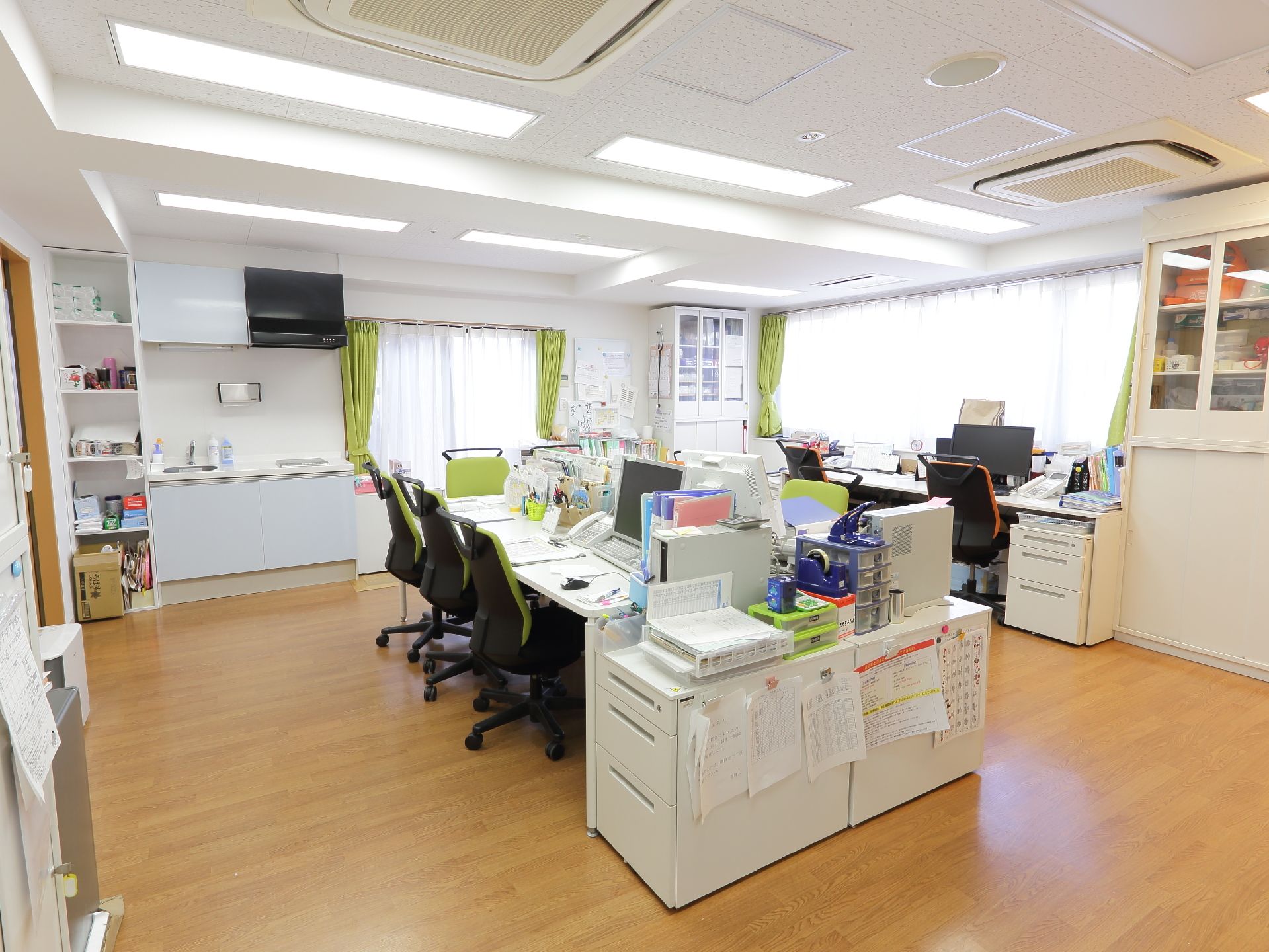 2面の窓から明るい光が差し込んでいる。事務机が向かい合う形で何台も置かれており様々な書類が置かれている。