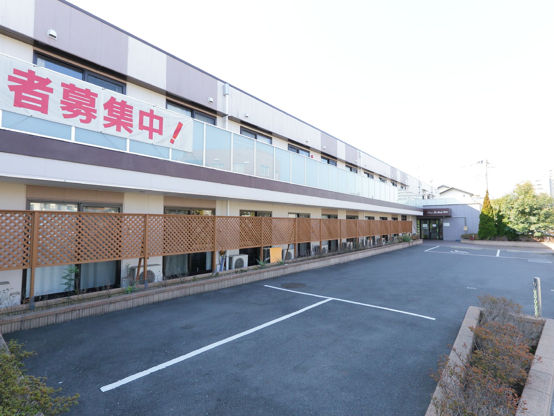 2階建ての細長い建物でベランダのついた施設になっている。木製のフェンス越しに舗装された駐車場が設備されている。