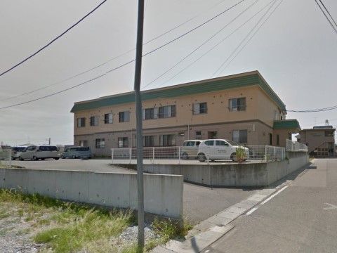 建物脇の道路から盛土をした場所に、ベージュの新築２階建の建物がある。屋根と玄関上のひさしは緑色でアクセントになっている。