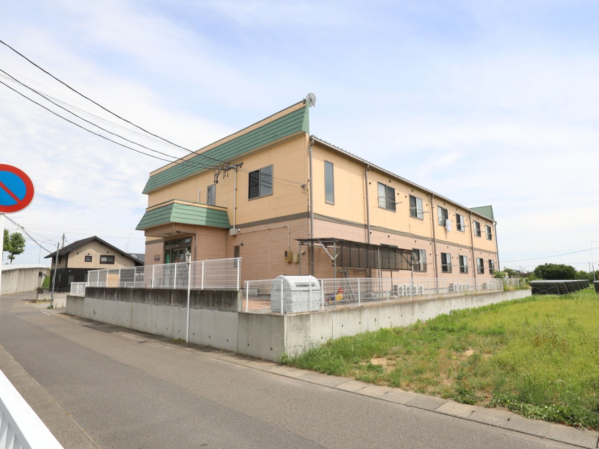 のどかな田畑に囲まれた2階建ての施設である。明るい色合いの外観。まわりには白色のフェンスが設置されている。