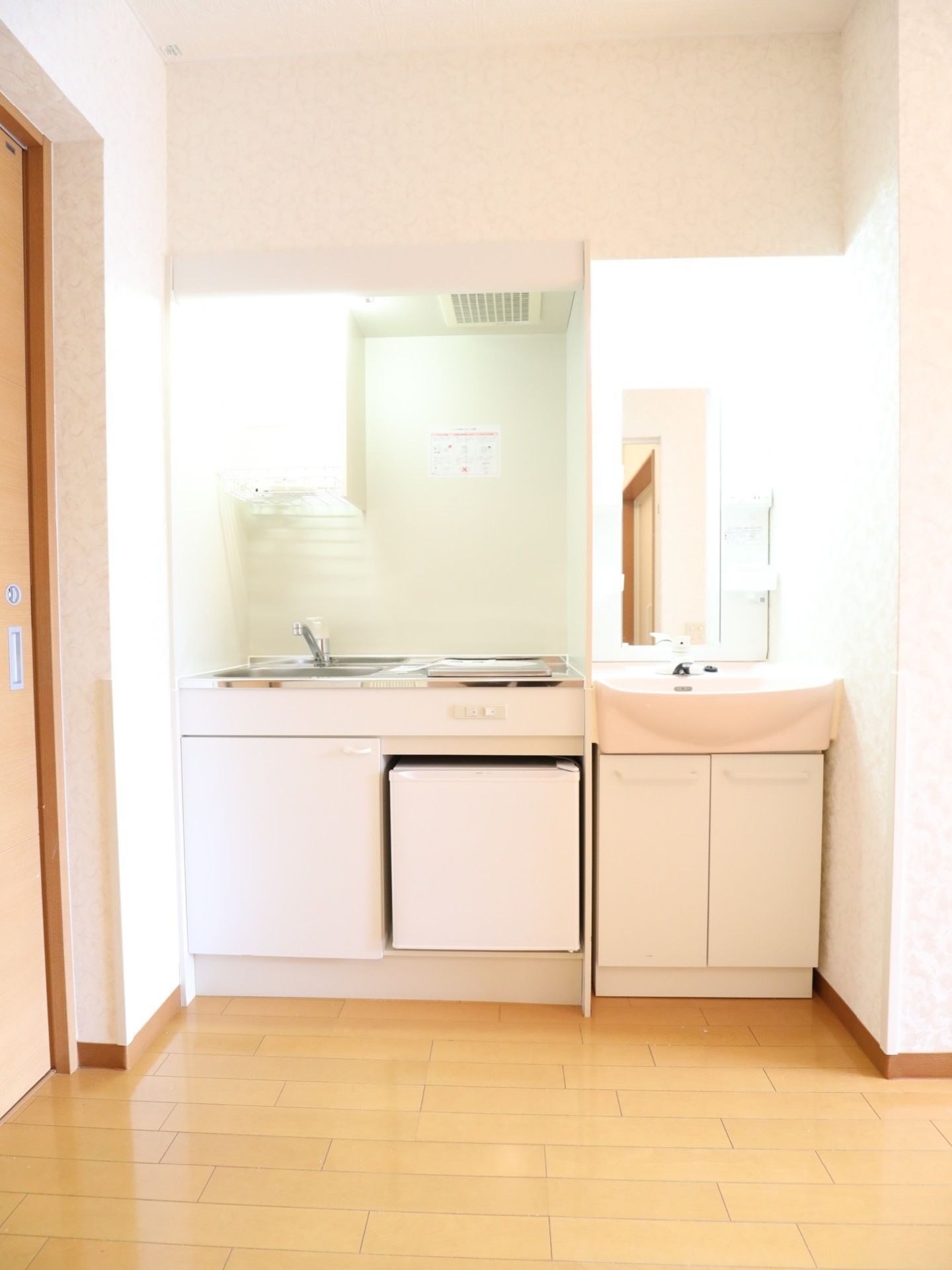 淡いピンク色の内装にクリーム色のフローリングの居室のすみに、ミニキッチンと洗面台が並んで設置されている。