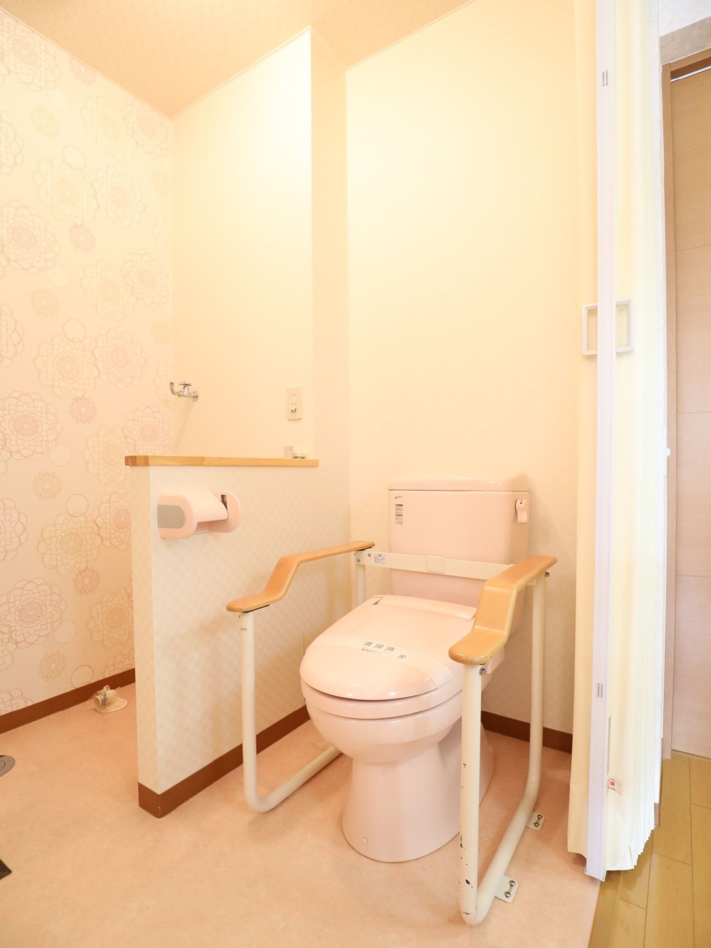 居室内にアコーディオンカーテンで仕切られた洋式トイレがある。便座の両横に手すりがあり、右手にペーパーホルダーがある。