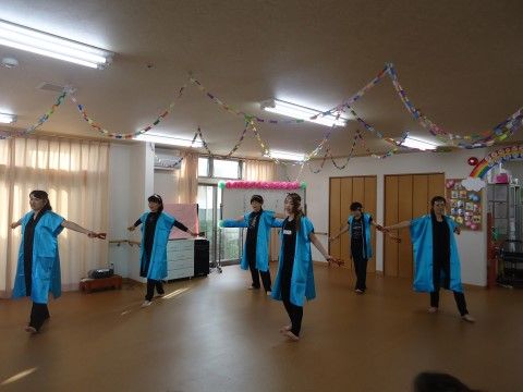 広い共用スペースで青い半袖の衣装を着用し、踊る６人のスタッフ