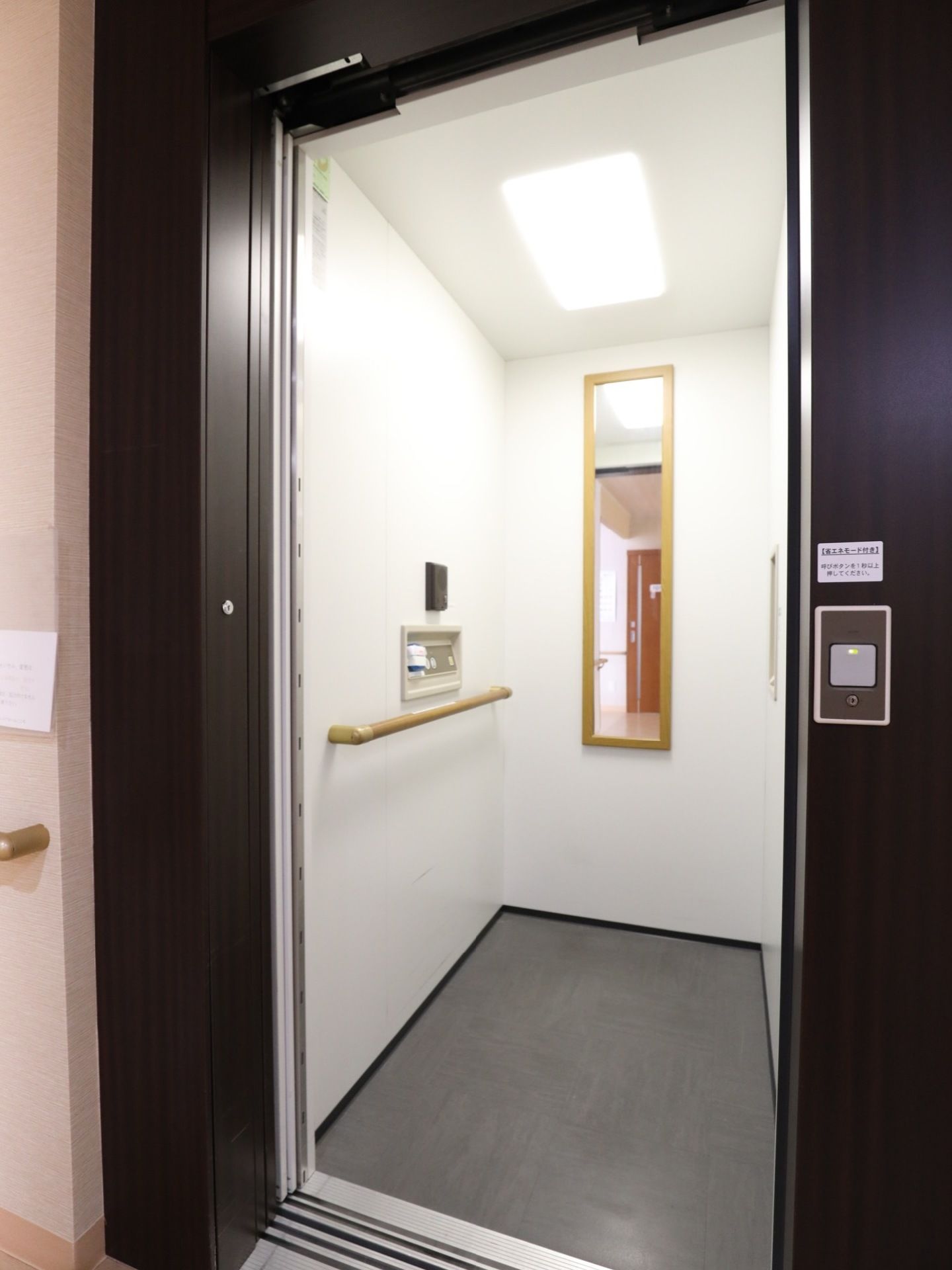 木目調のドアのエレベーター。ボタンは車いすの方も使用しやすいように低めに設置されている。中には姿見、手すり、呼出ボタンがついている。