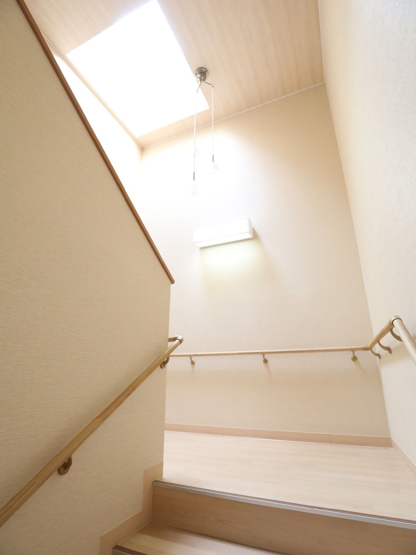 上から陽光が差し込んでいる明るい踊り場。木目調の床と天井。壁には入居者様の安全のために手すりがついている。