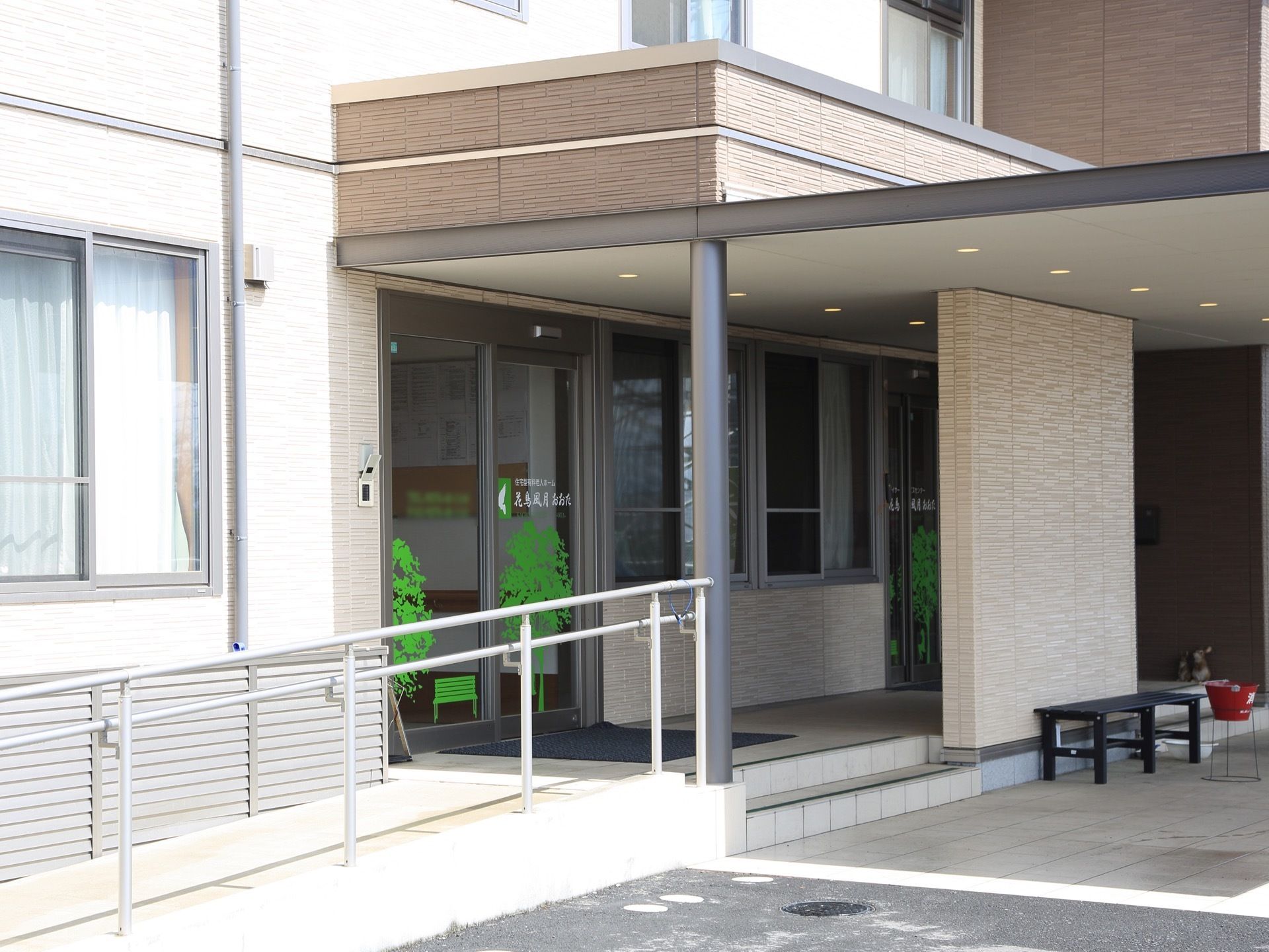 外壁が白い建物である。玄関前はひさしが伸びており、喫煙スペースが設けられている。喫煙スペースにはベンチと灰皿が置かれている。