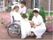 中庭の一角に看護師２人と車椅子の入居者様がいる。芝生に繋がる植え込みには紫色の花が咲いている。３人とも笑顔で話をしている。