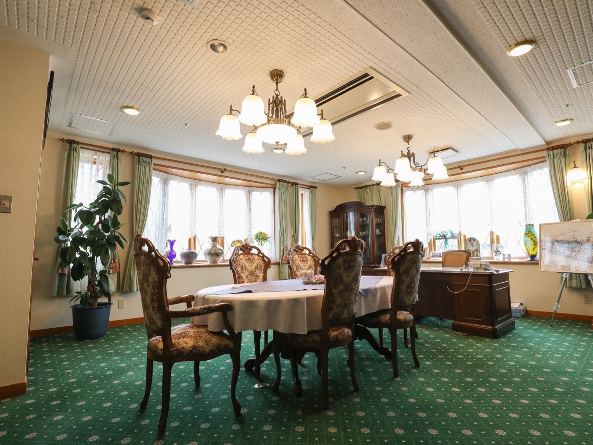 緑色に白色の模様が入った絨毯が敷かれている。部屋の中央にはテーブルと5脚の椅子が並べられている。窓際には複数の花瓶が並べられている。
