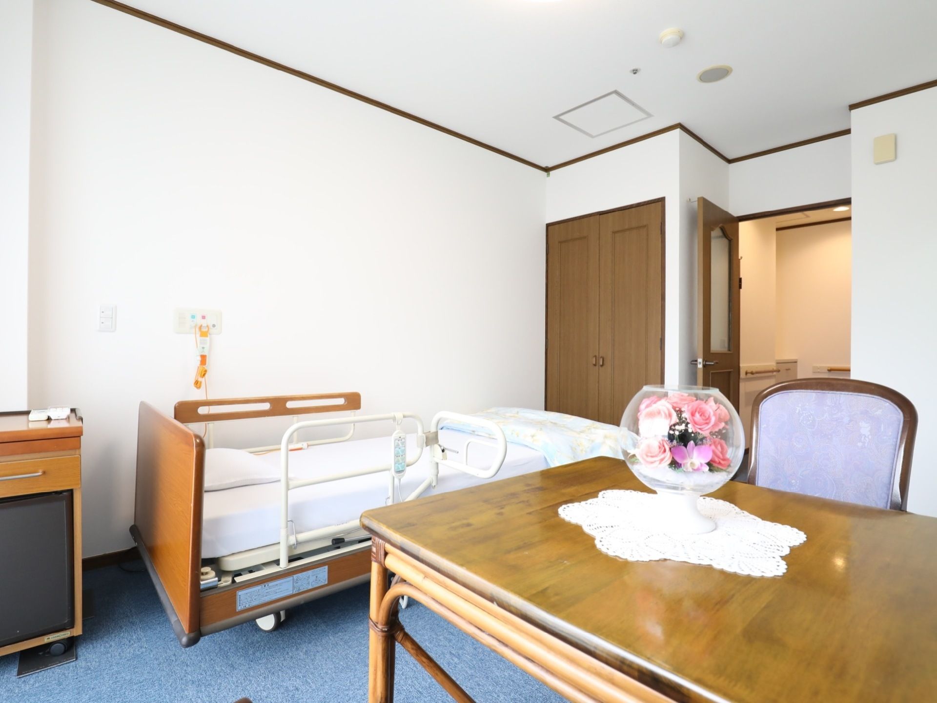 居室には柵付きのベッドが用意されている。テーブルや椅子もあり、テーブルの上にはピンク色の花が飾られている。