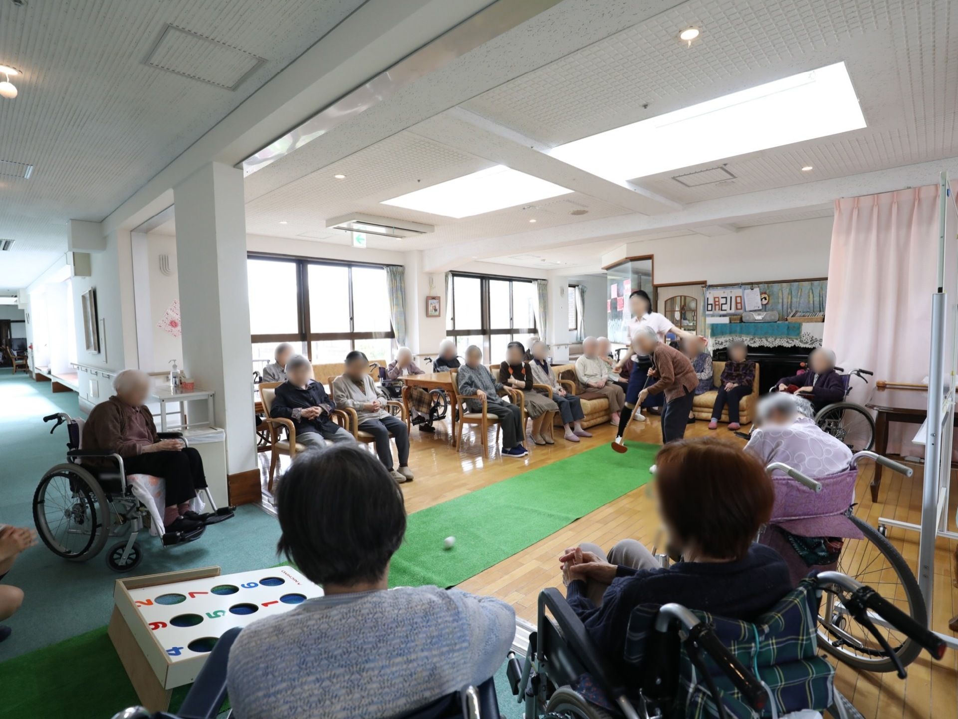 緑色のパターマットが部屋の中央に敷かれている。スタッフの近くで1人の入居者様がゴルフをしている。周りには他の入居様が見ている。