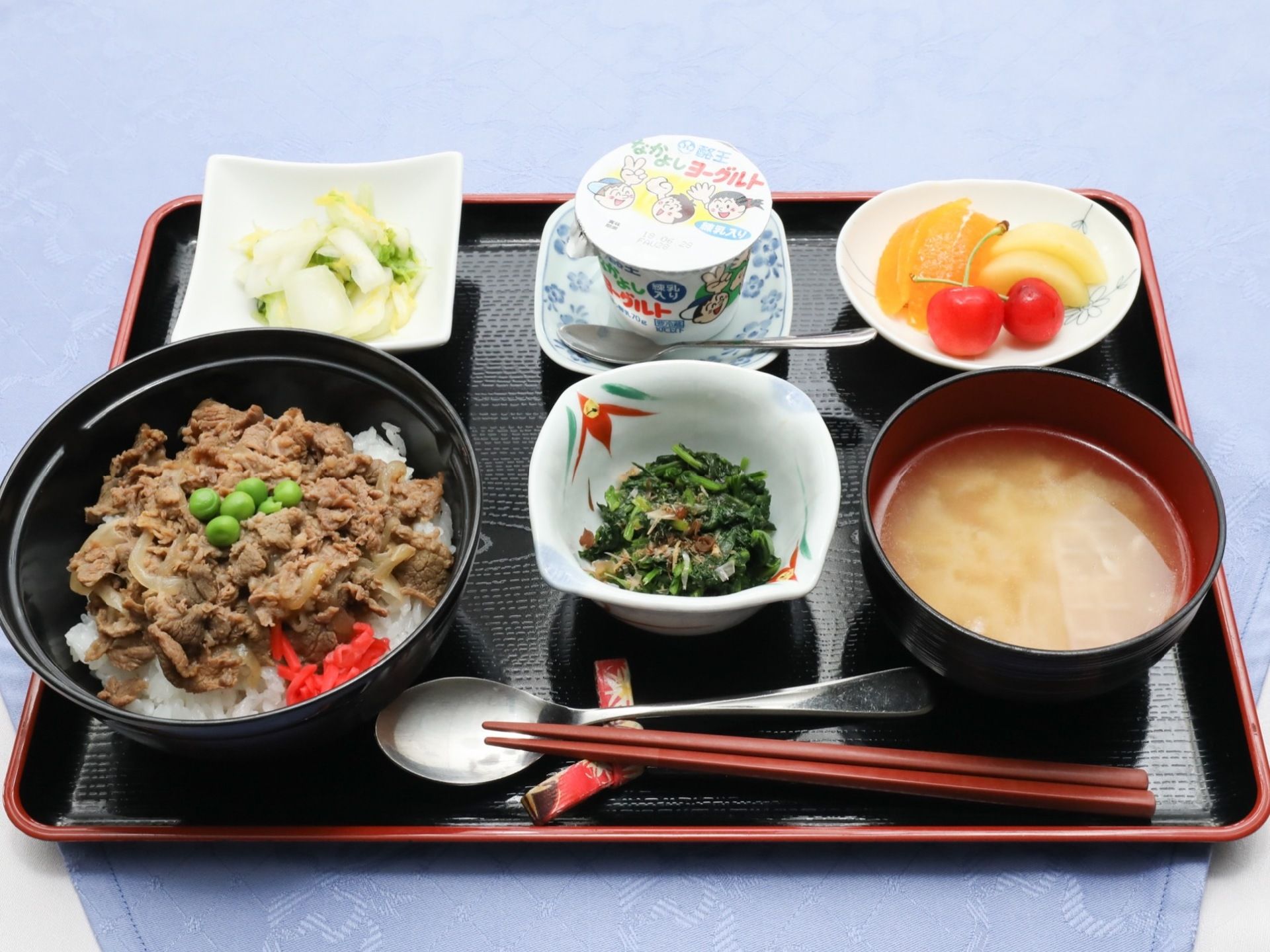 赤く縁取りされたおぼんの上には食事が配膳されている。メインの牛丼、白菜のお漬物、ほうれん草のおひたし、お味噌汁、フルーツ、ヨーグルトである。