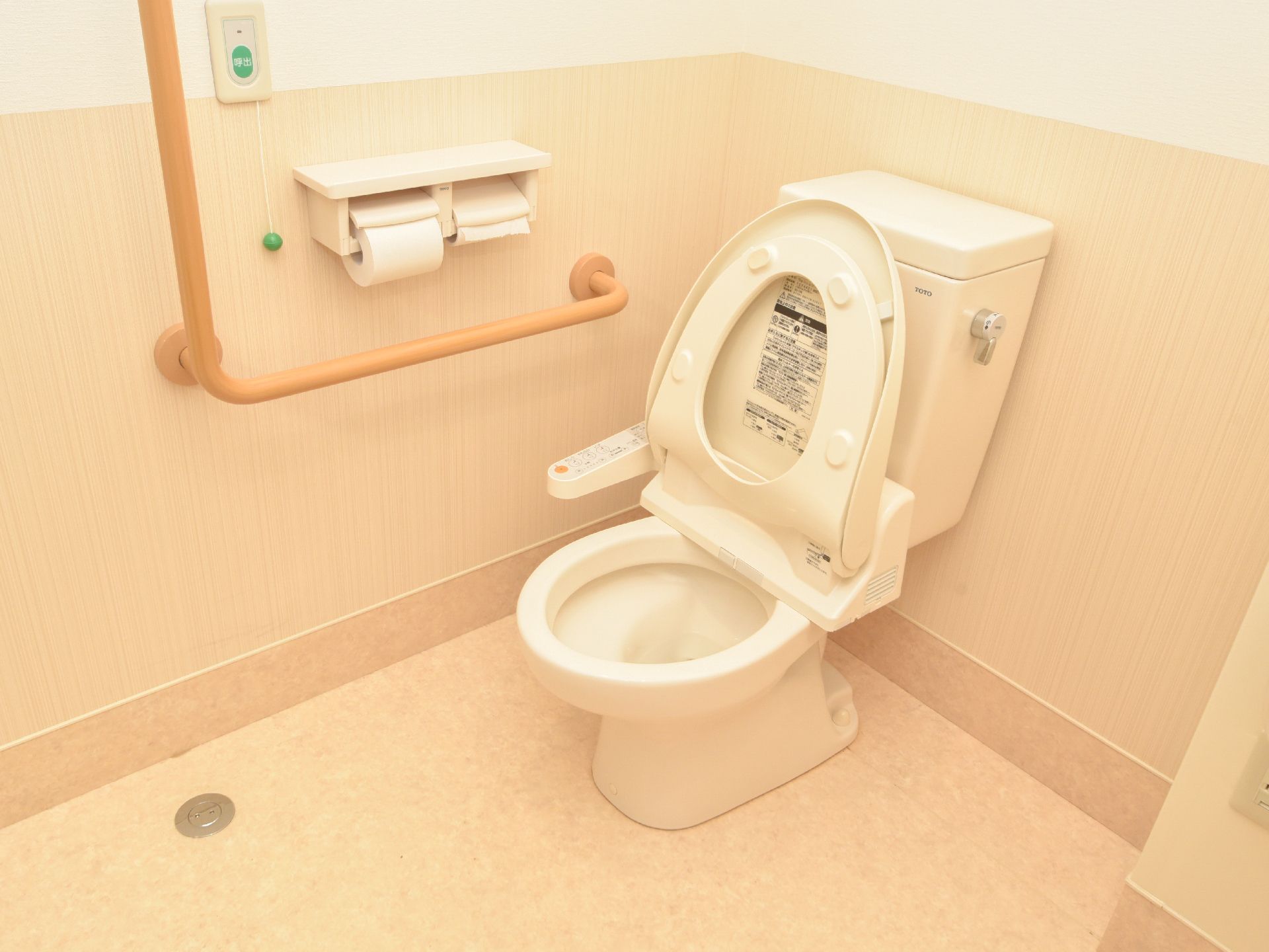 トイレの個室には、様式の便器の設置がある。便器は温水洗浄便座付きになっている。右の壁にはL型の手すりが設置されている。