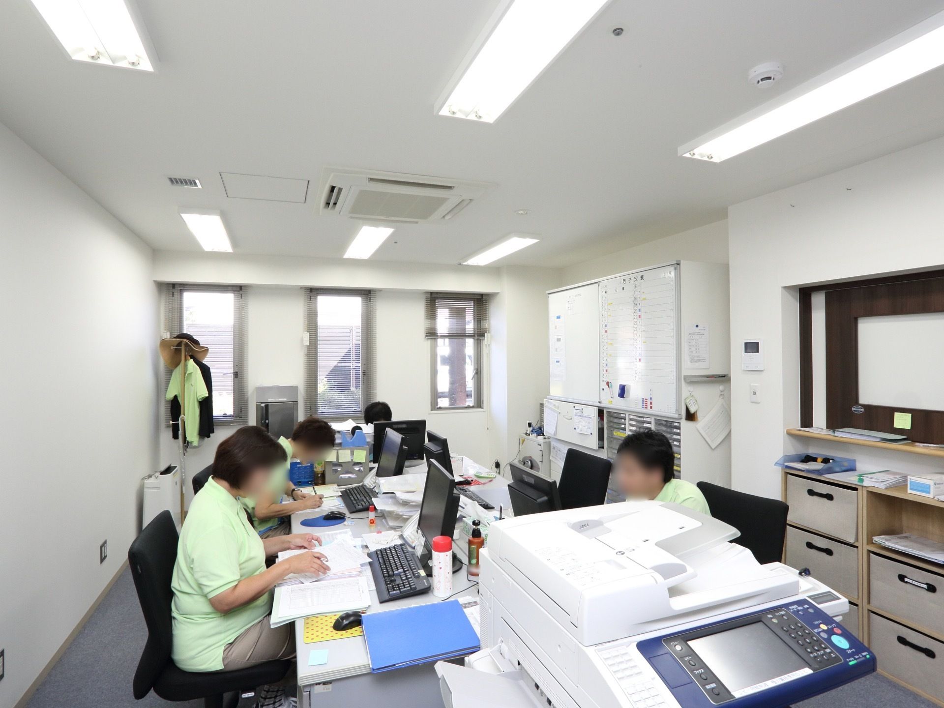 数名のスタッフが、デスクに座って事務作業をしている。書類棚やホワイトボードなどが置かれ、室内は整理整頓されている。