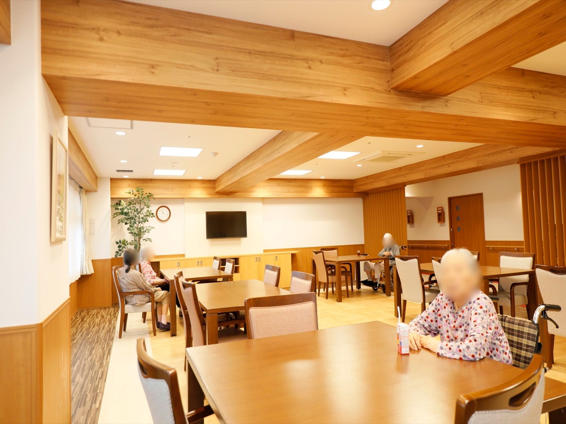 木目調デザインのテーブルと椅子が、数多く並べられている。手前のテーブルでは、入居者様がジュースを飲みながらくつろいでいる。