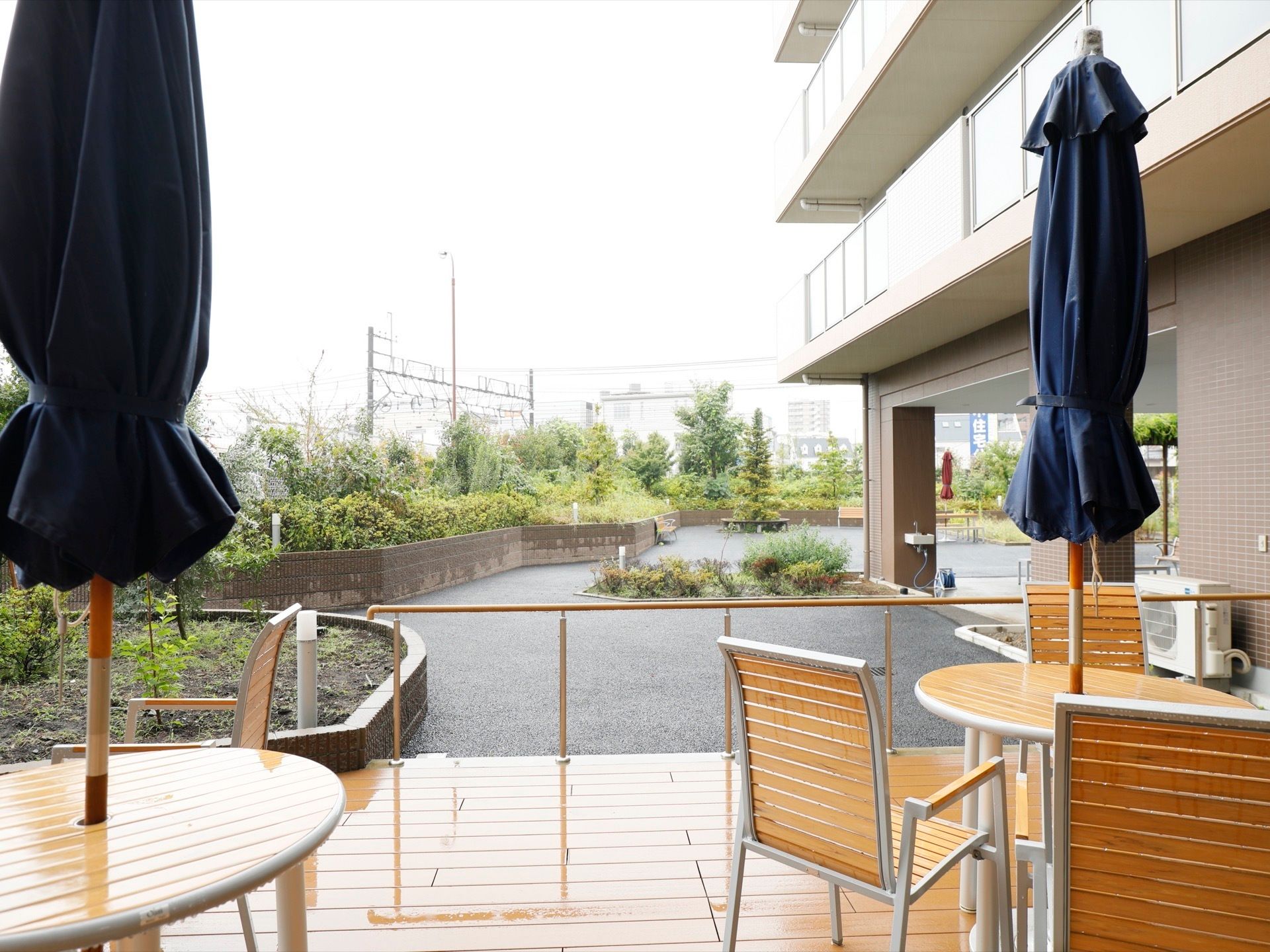 ガーデンテーブルの真ん中に、紺色のパラソルが取りつけられている。柵の向こうには植え込みがあり、草木が茂っている。