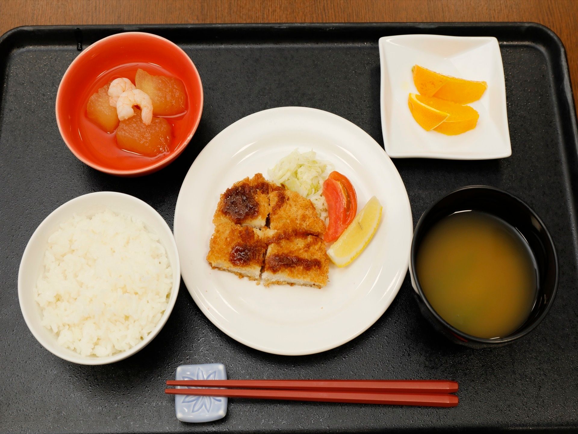 ご飯、お味噌汁、とんかつ、大根とエビの煮物、デザートのオレンジが、黒いトレイに、バランス良く並べられている。