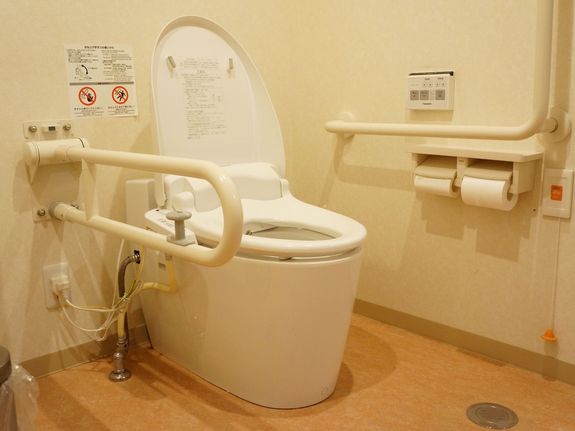 温水洗浄便座が設置され、壁にはL型や可動式の手すり、操作パネル、トイレットペーパーが備えてある。後ろの壁には使用上の注意書きが貼ってある。