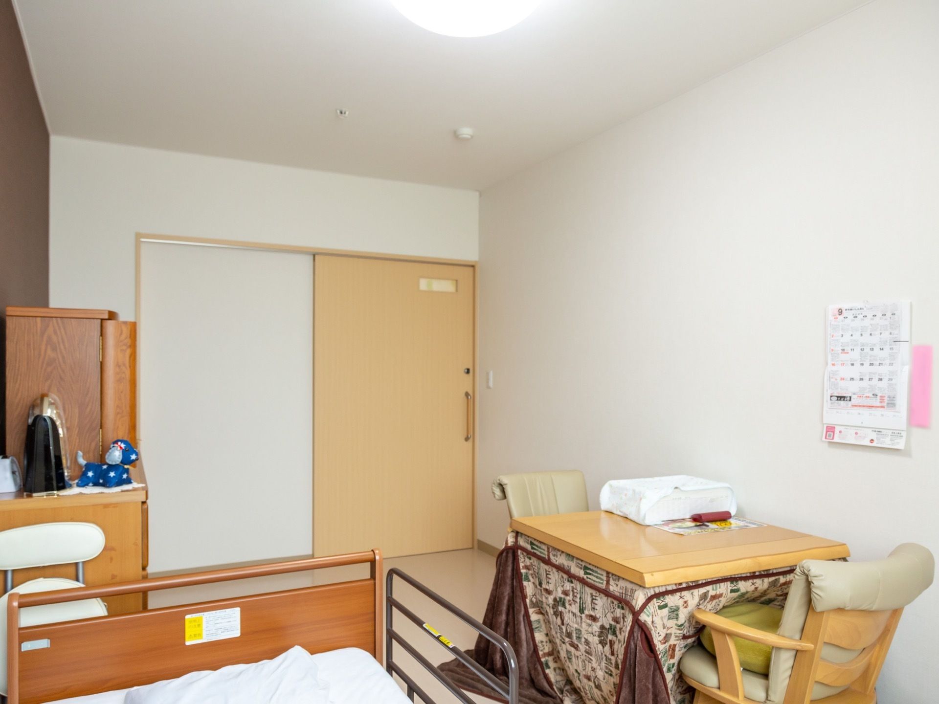 明るい色合いの居室である。扉の上部に横長の明かり窓を設置。ベッドやこたつ、収納家具などが配置されている。