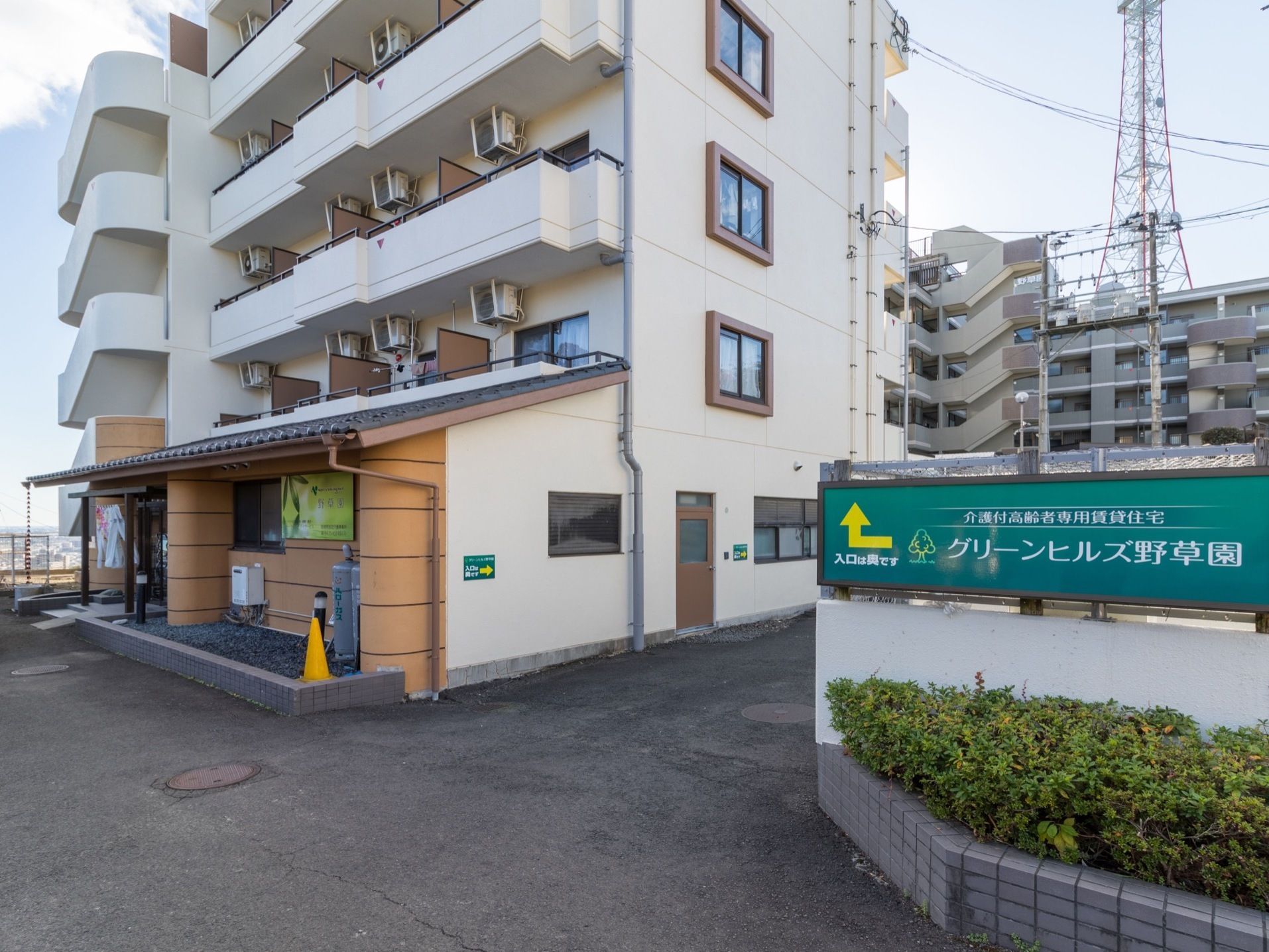 5階建ての建物で、1階部分には瓦の屋根がついている。右側に花壇があり、その上に施設名の書かれたグリーンの看板が設置されている。