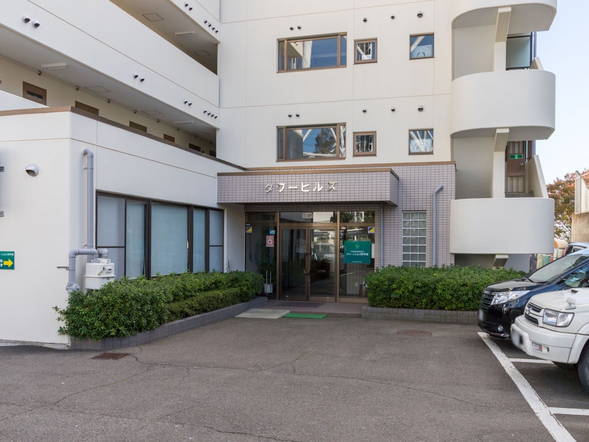 白い建物で玄関部分はベージュのレンガ調になっている。玄関の近くには花壇があり、建物の右側には駐車スペースがある。