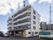 5階建ての白い建物で、2階以上にはベランダがついている。周りにはマンションや電波塔などが建っている。