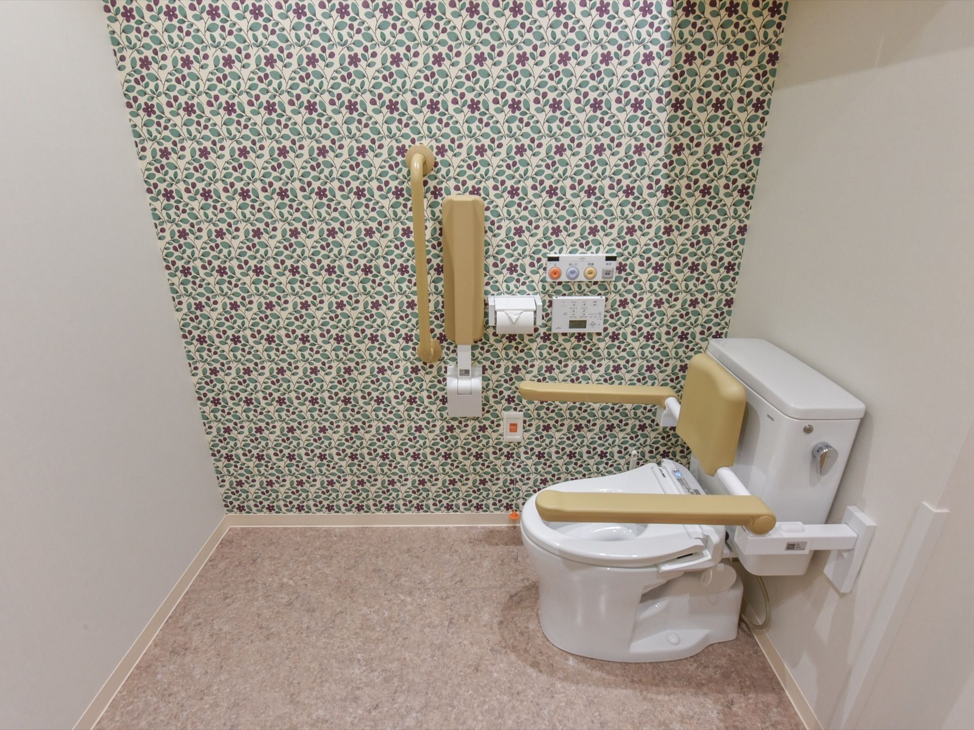 奥の壁が花柄で、スペースに余裕のあるトイレである。背あてや可動式の手すりの付いた、温水洗浄機能付き便座である。