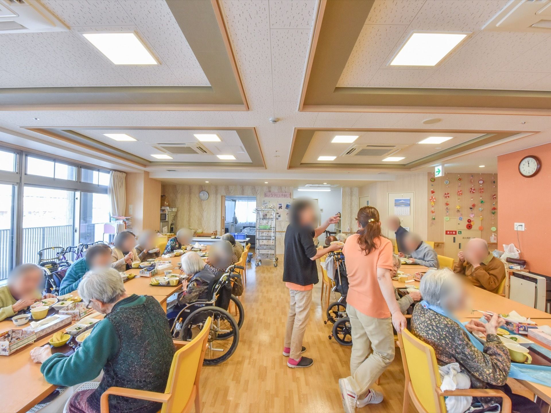 ２列に長く並べられたテーブルで、入居者様が食事をしている。左側は大きな窓になっており、バルコニーの柵が見える。