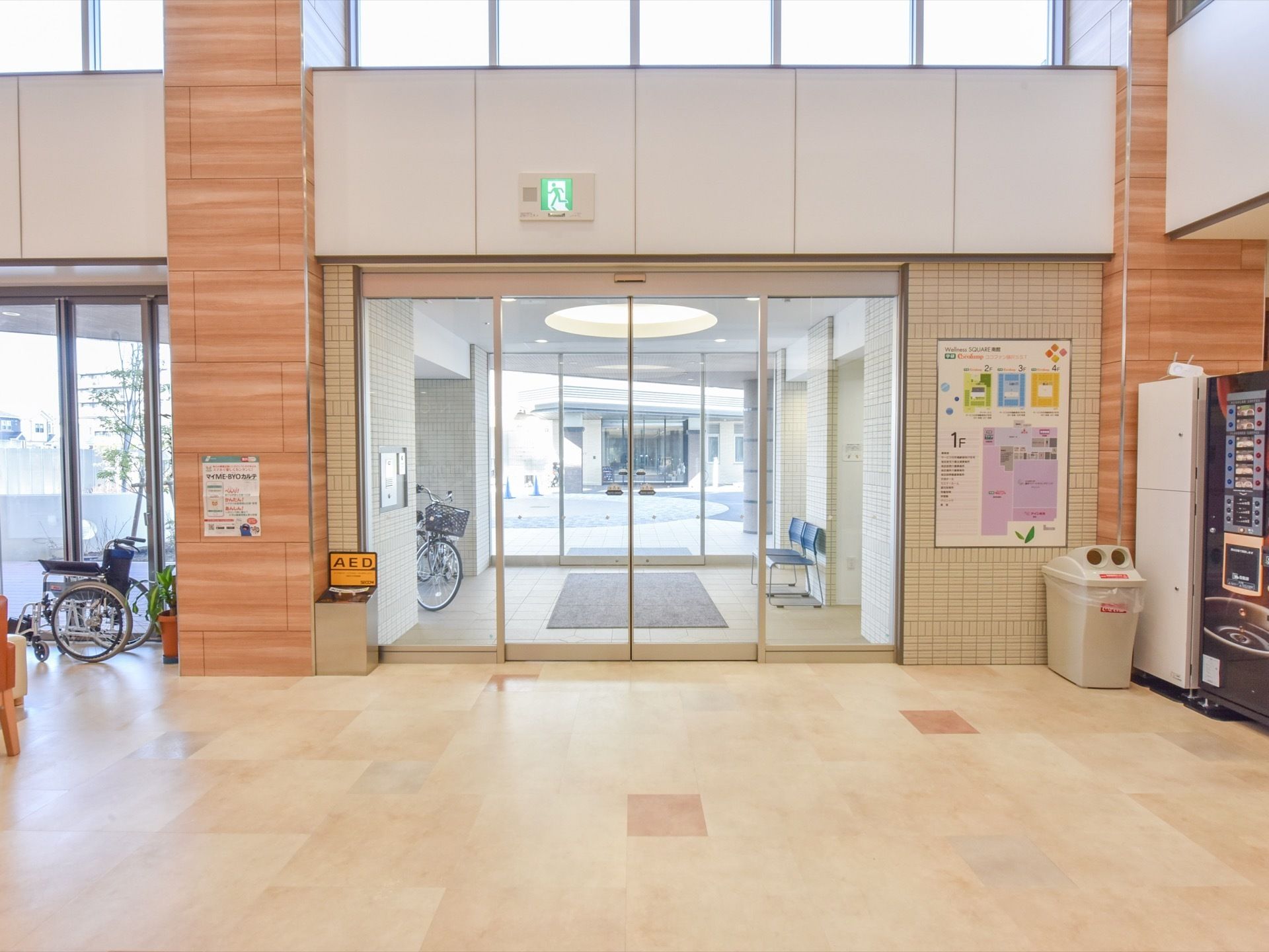 玄関のドアは、透明なガラスの大きな自動ドアである。風除室には椅子や自転車が置かれている。玄関内には自動販売機が置かれている。