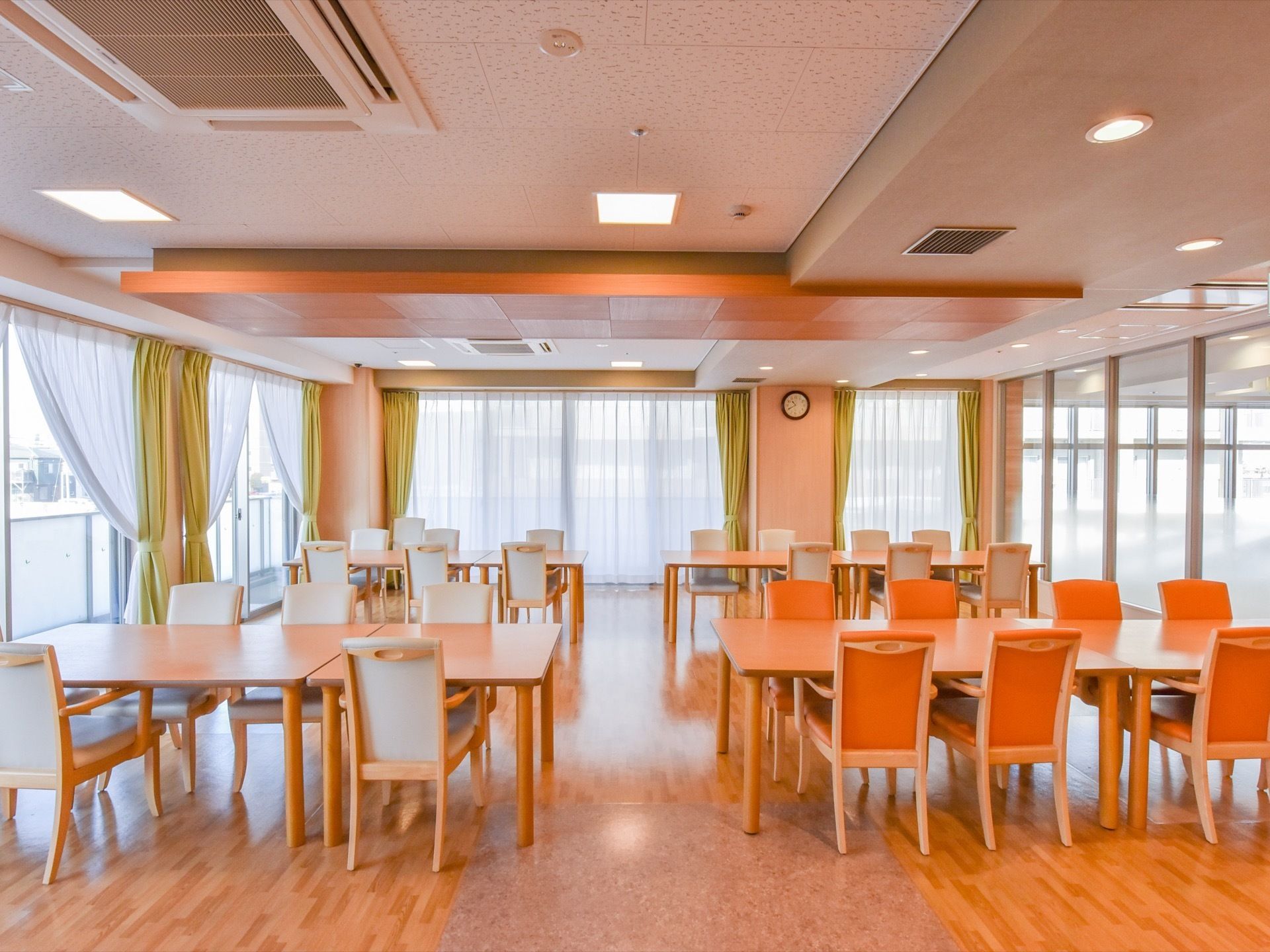 窓に囲まれた明るい食堂に、ダイニングテーブルが余裕を持って置かれている。椅子は座面が白色のものとオレンジ色のものがある。