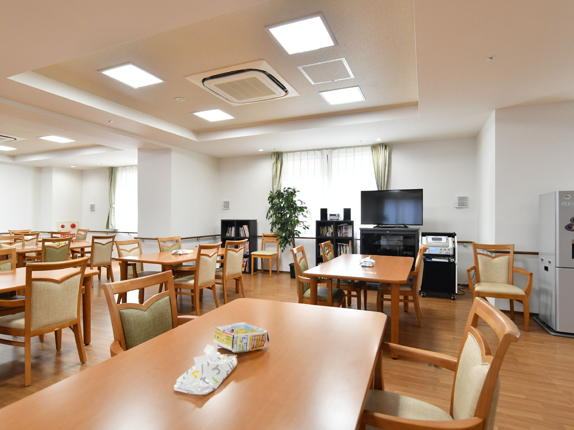 正面奥にはテレビが置かれている。その左側には棚があり、その左隣には観葉植物が置かれている。沢山のテーブルと椅子がある。