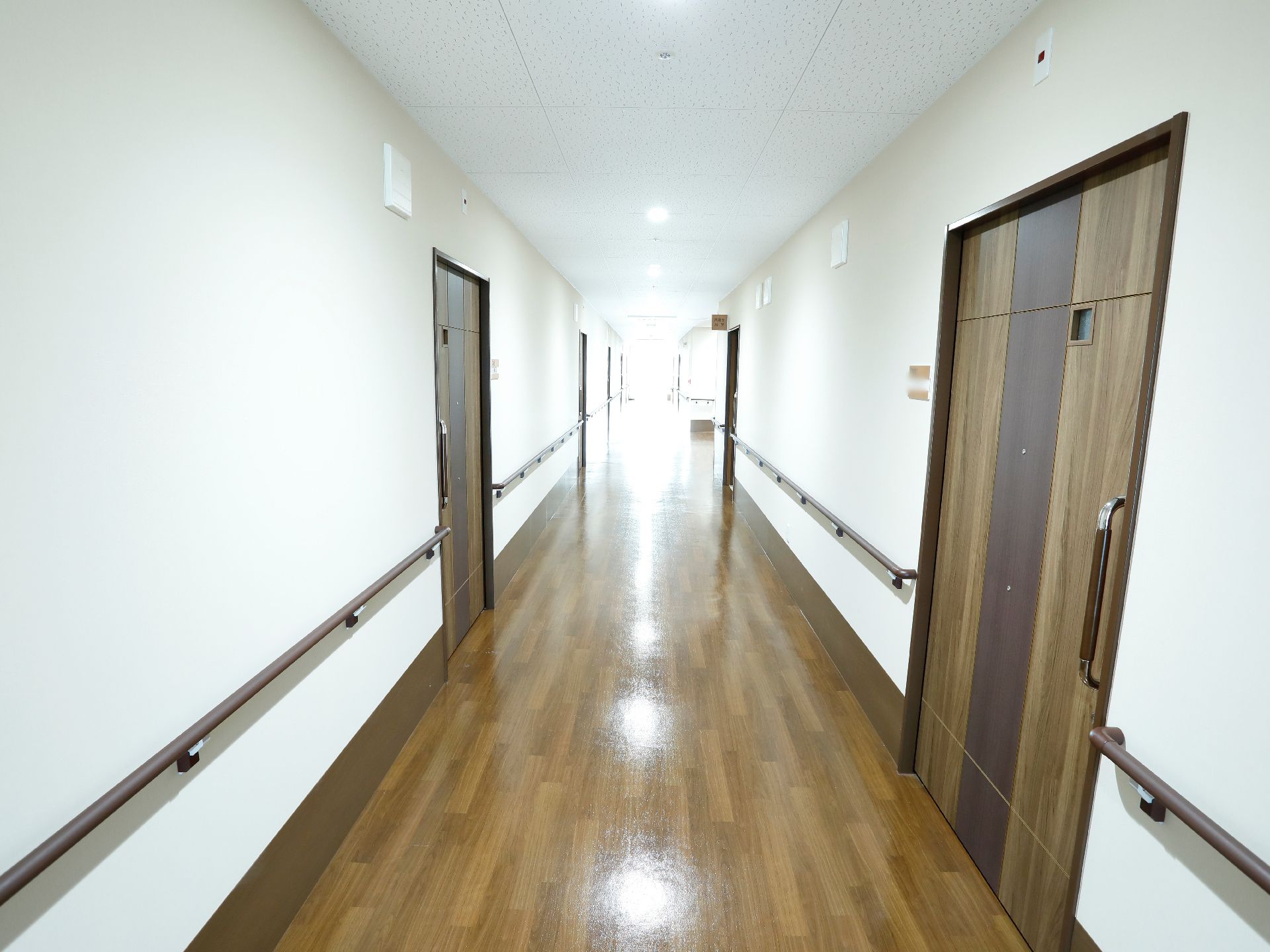 白い壁の廊下の両脇には、濃い茶色の個室のドアが並ぶ。廊下の壁には横型の手すりが、個室のドアには縦型の手すりが備えられている。