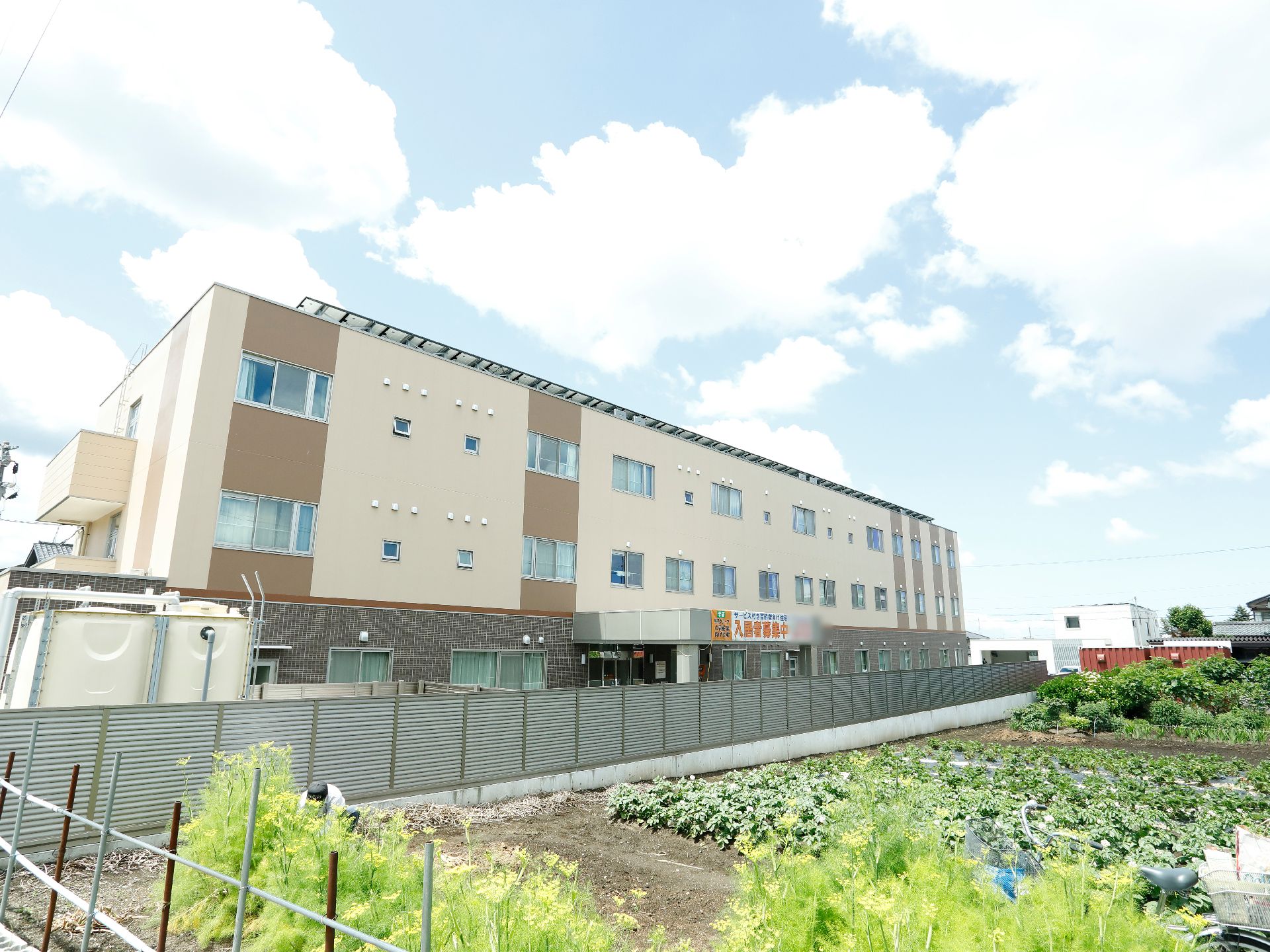 左側には建物があり、その手前には貯水槽が置かれている。右側には畑がある。建物と畑の間にはフェンスが設置されている。