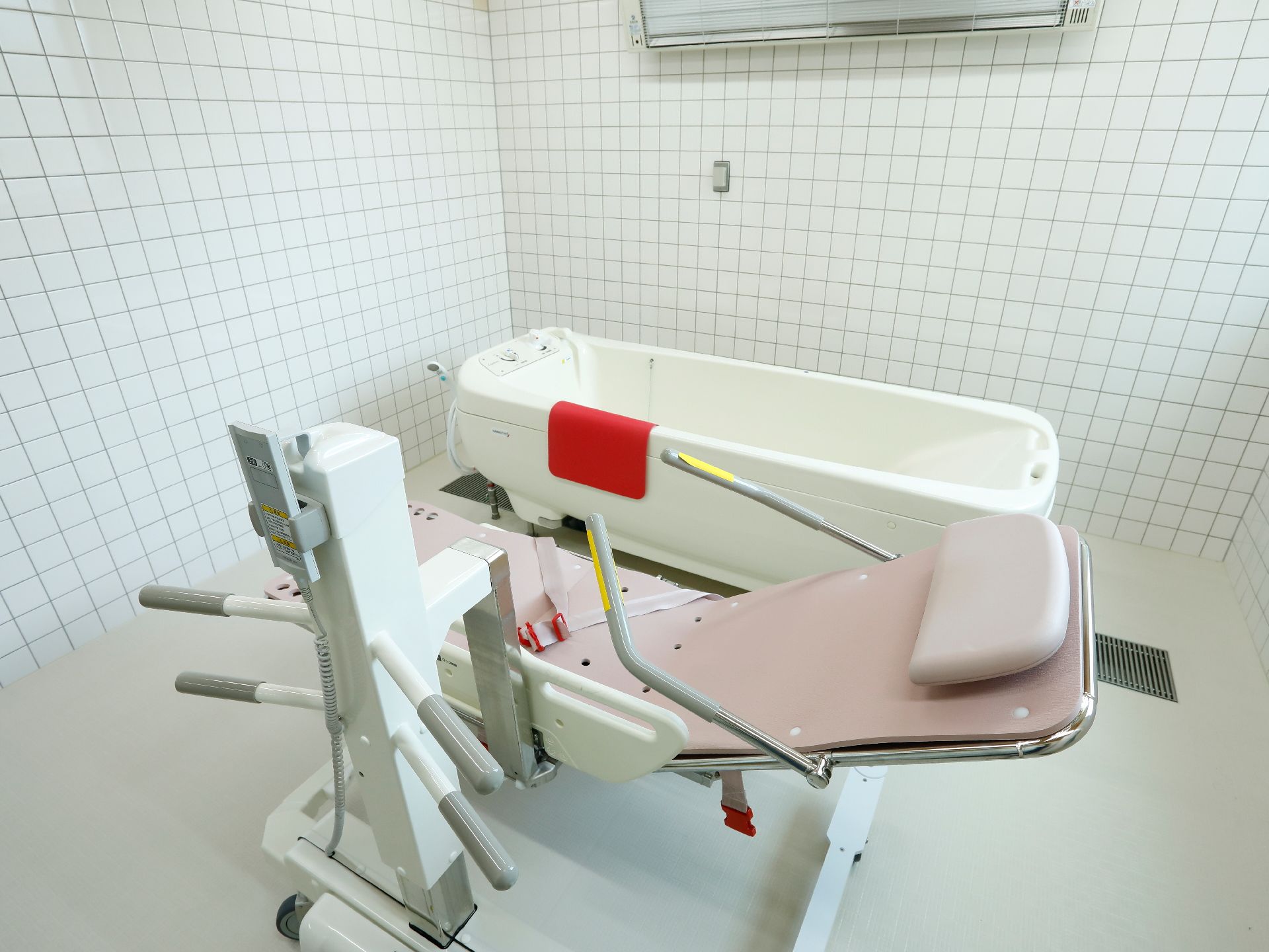 左側には介護用入浴機器がある。右側には浴槽がある。浴槽には赤いタオルの様なものが掛けられている。床は白色になっている。