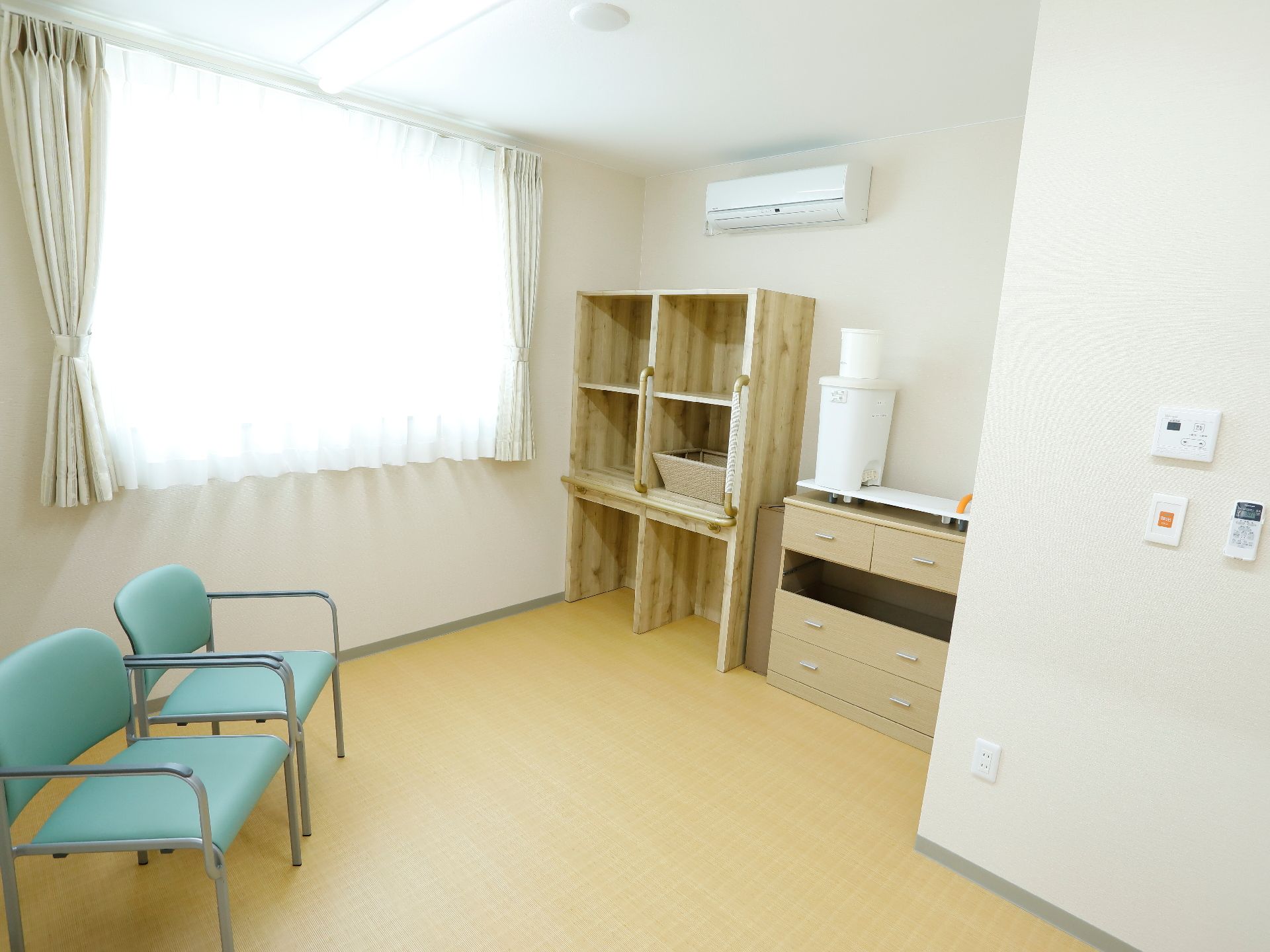 左側には窓があり、その前には椅子が2脚ある。右側にはエアコンが付いていて、下側には棚と箪笥がある。右側の壁には緊急呼び出し装置がある。
