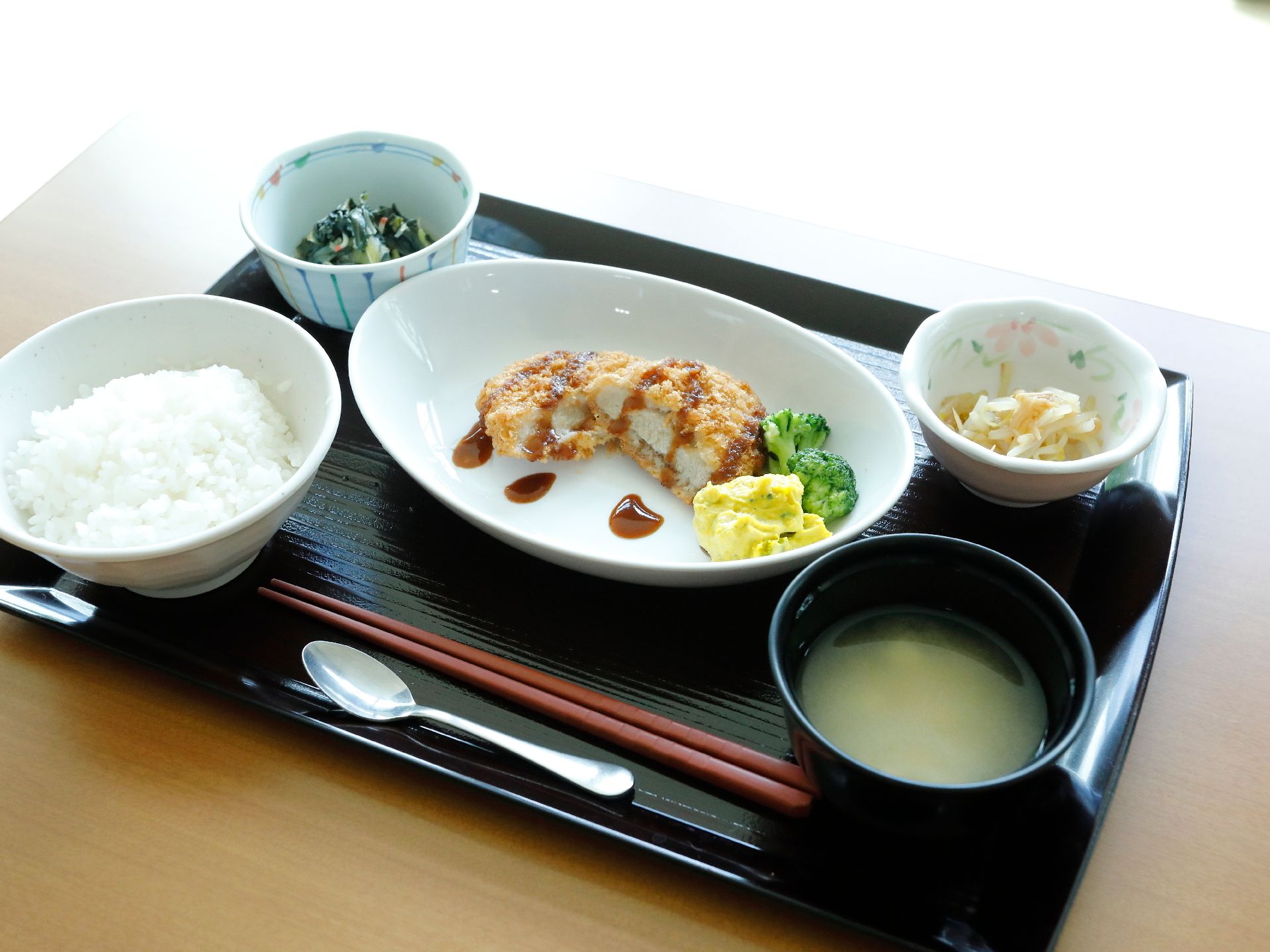 黒いお盆の上に箸とスプーンが置かれている。お盆にはご飯と味噌汁があり、その他おかずが3品乗せられている。