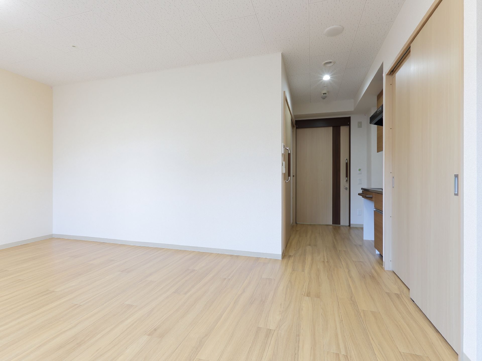 右奥には扉があり、近くにはキッチンスペースがある。床はフローリングになっている。壁は白色に塗られている。