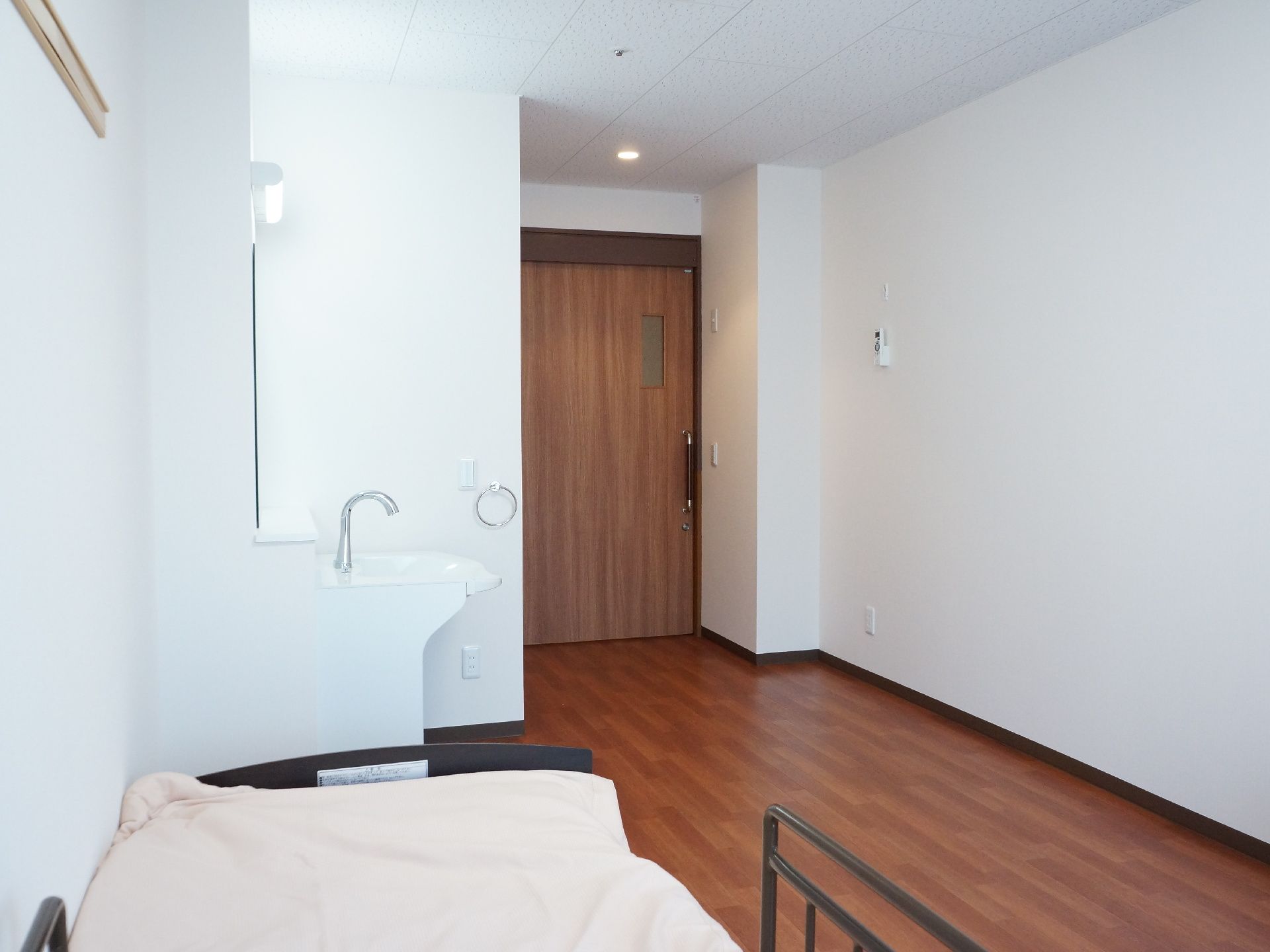 白とこげ茶色が基調となっている部屋。横開きの扉で、入口付近には、洗面台、奥にはベッドが設置されている。