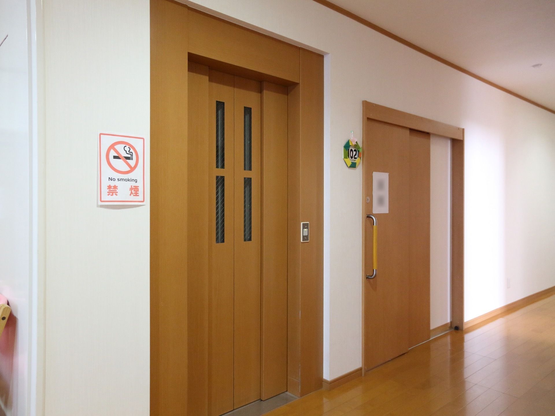 白い壁で清潔感のある廊下になっている。扉がベージュ色のエレベーターがあり、その隣には禁煙のポスターが貼られている。