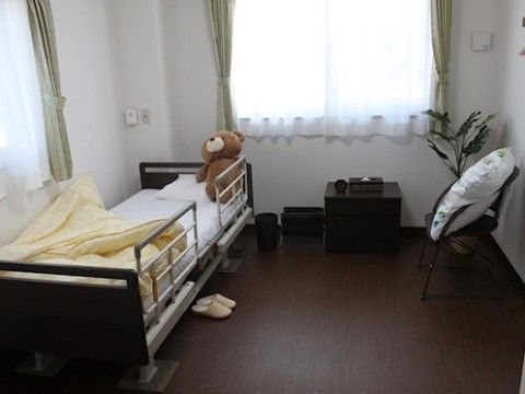 白い壁にダークブラウンの床の室内。二か所に窓がある。窓際にはベッド、椅子、観葉植物などが置かれている。