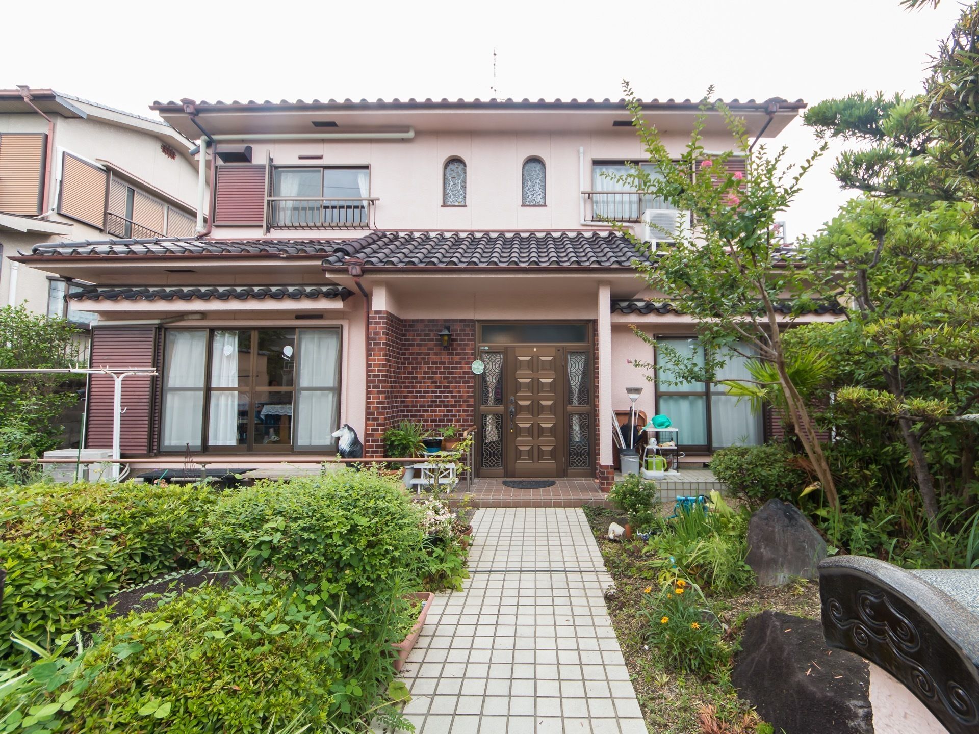 ベージュを基調とした外壁で、屋根は黒の瓦の屋根である。白のタイルの道のわきには、木がたくさん植えられている。