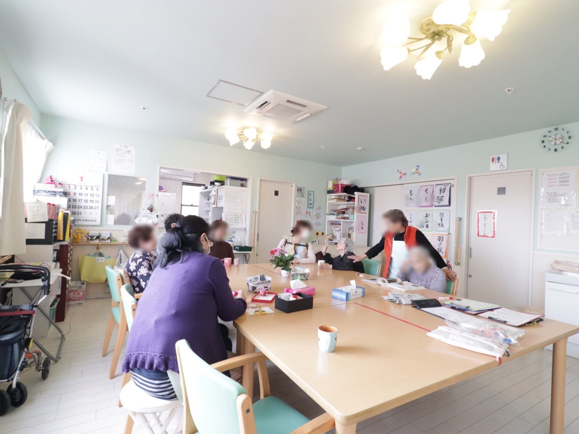広いテーブルのまわりをかこむようにスタッフと入居者様が集まり、レクリエーションをしている。そばにトイレも設置されている。